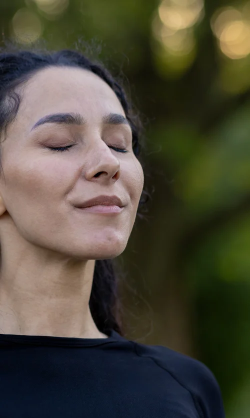Woman standing outdoors with eyes closed, practicing calm, controlled breathing with relaxed posture